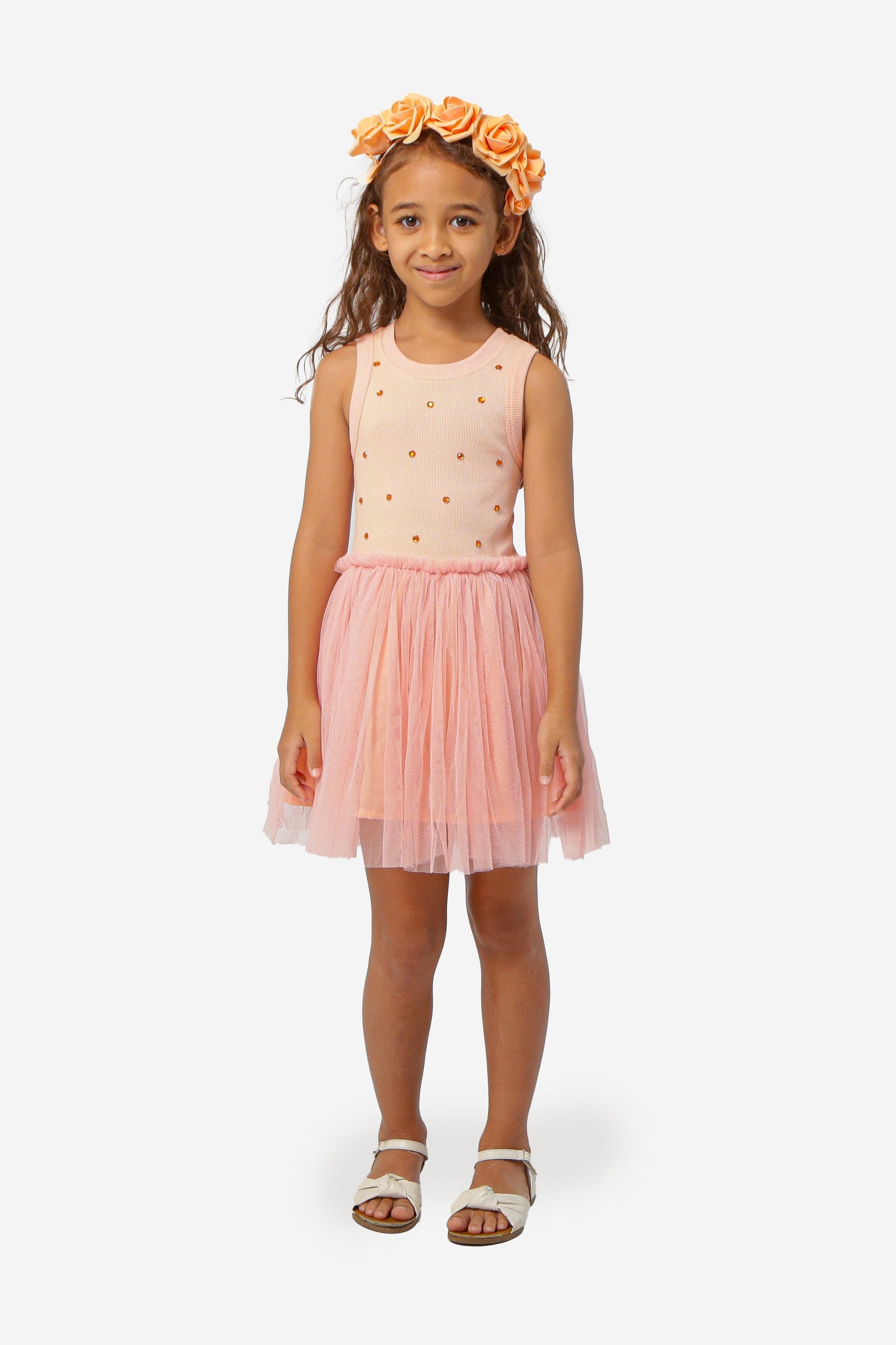Young girl wearing a peach dress with a sequin dot top, tulle bottom, and a flower crown on a white background