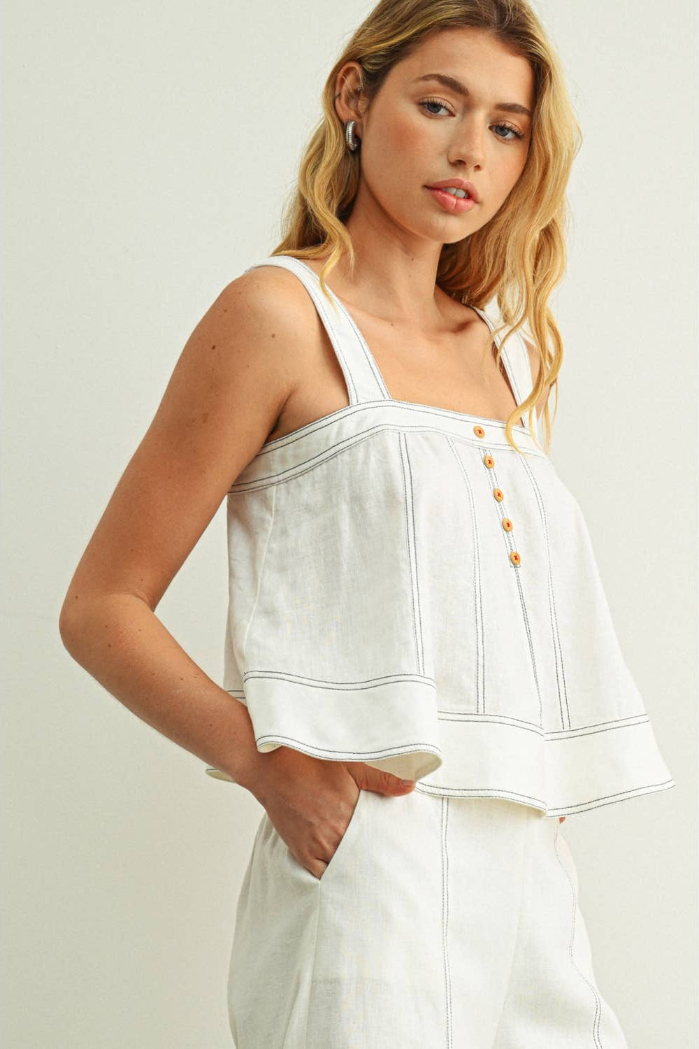 A woman wearing a white sleeveless cropped top with thin straps, vertical seam detailing, and a row of gold buttons down the front, paired with matching white pants, set against a plain light gray studio background.