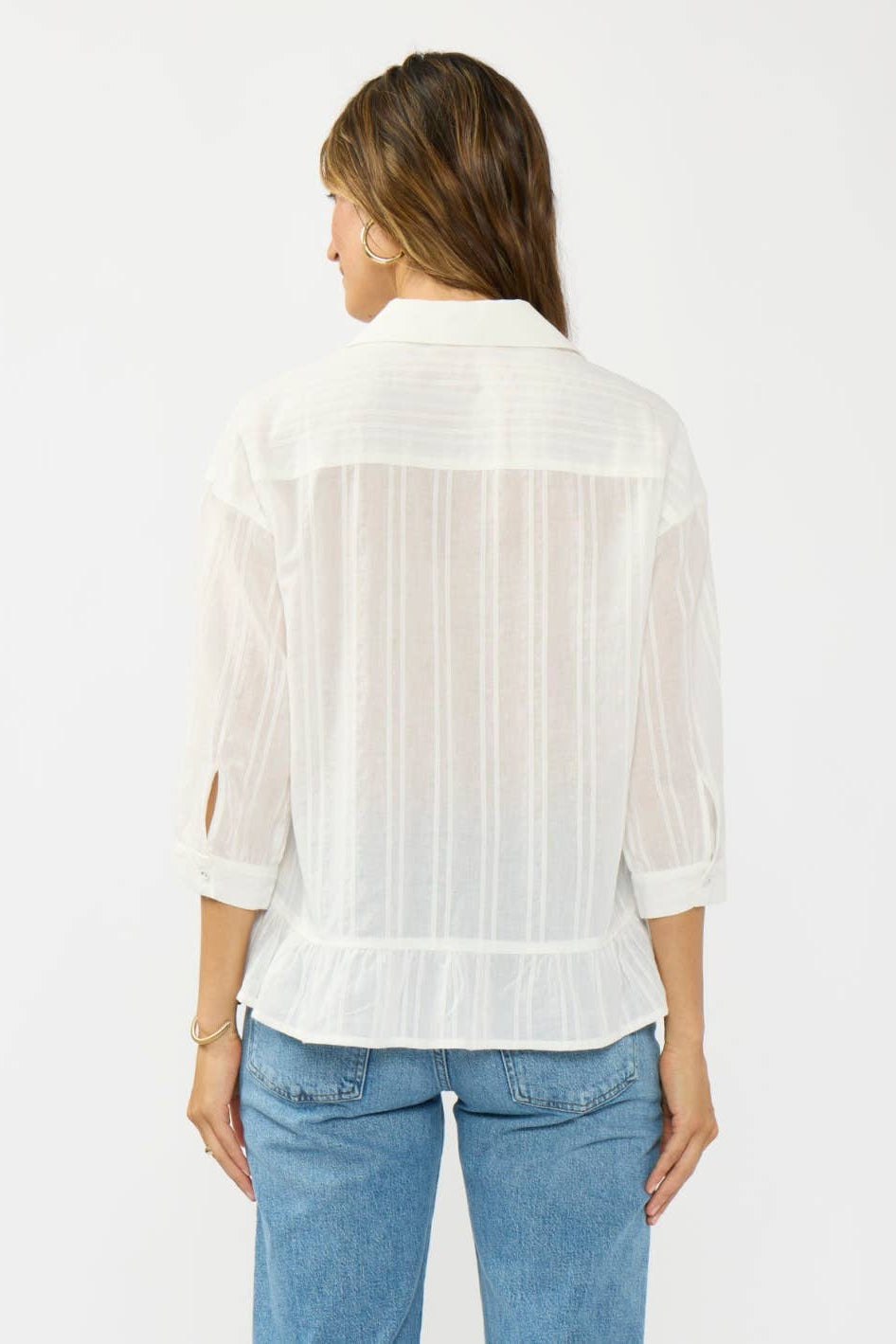 A woman viewed from the back wearing a white sheer long-sleeve blouse with vertical texture details and light blue jeans, set against a plain light gray studio background.
