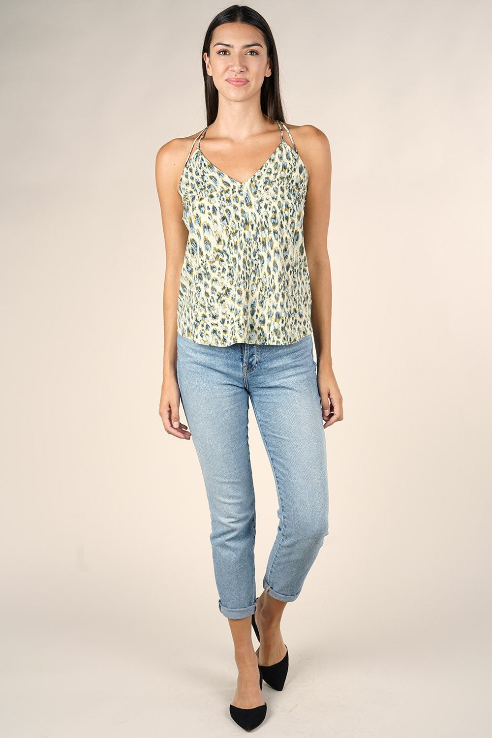 A woman modeling a sleeveless V-neck camisole top with a safari/lagoon pattern, paired with light blue cropped jeans and black heels, against a solid beige studio background.