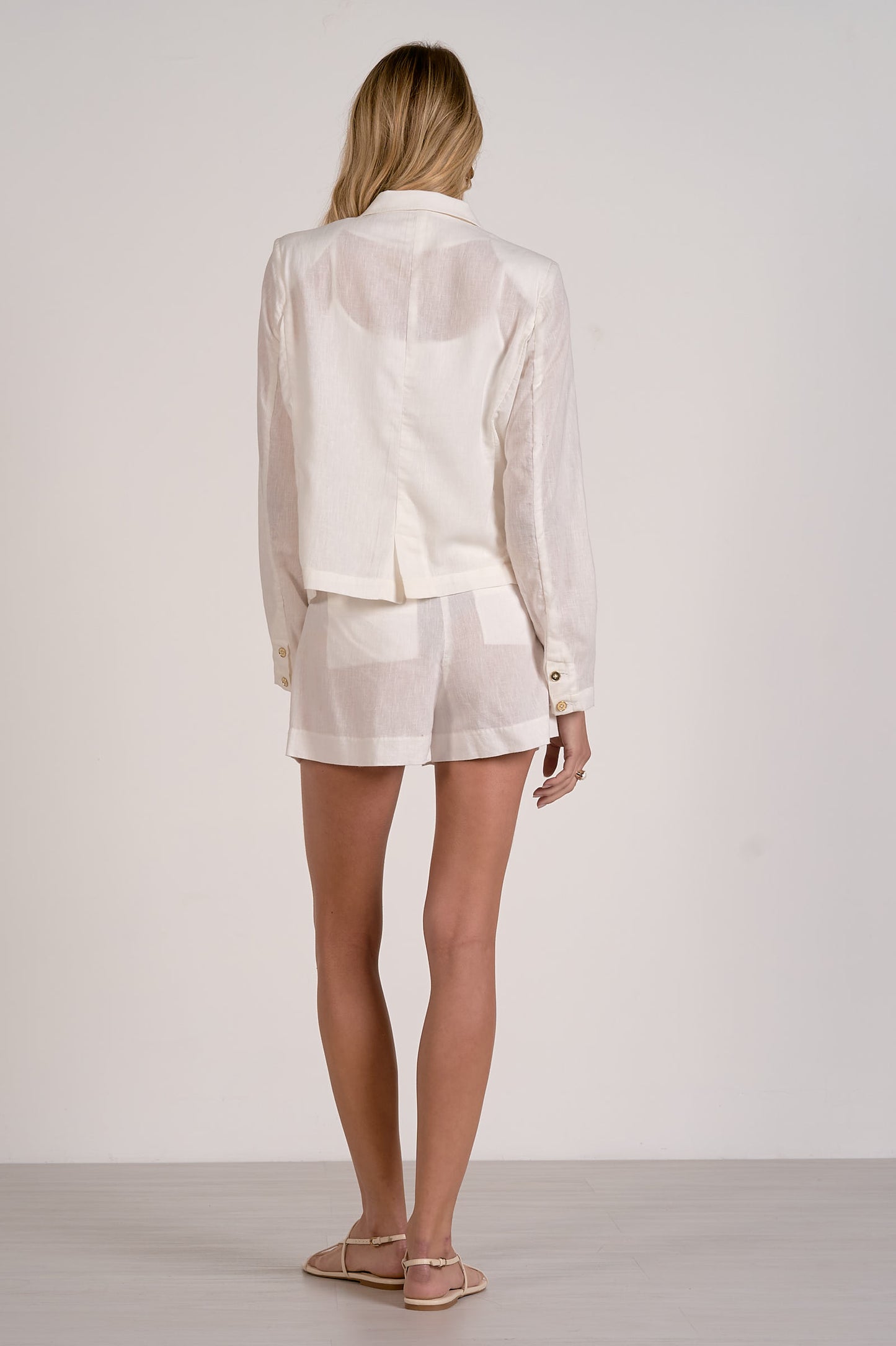 Back view of a woman wearing a white blazer and white shorts with sandals on a plain background.