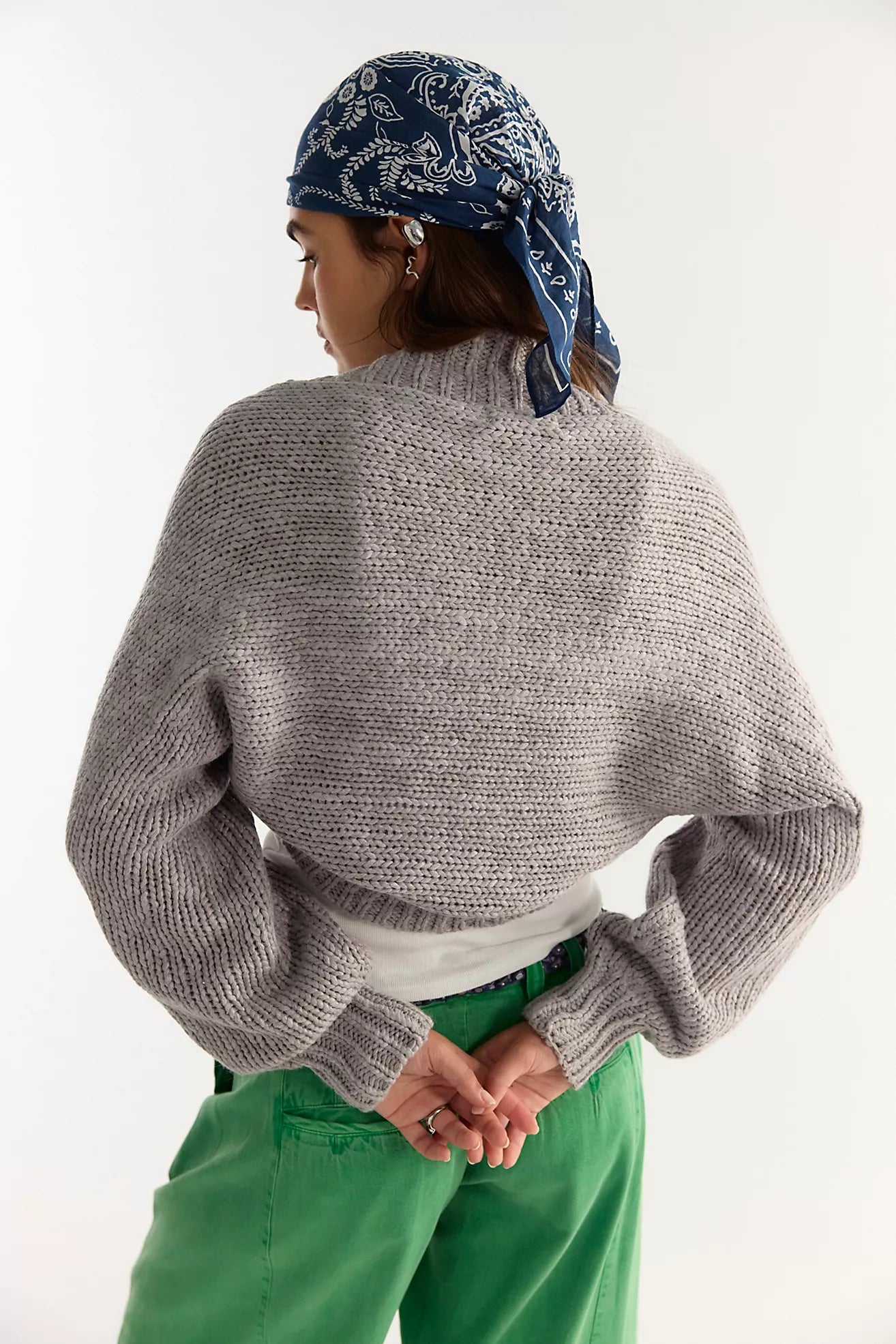 A person shown from the back wearing a chunky gray knit mock-neck sweater, a blue paisley bandana, and green pants against a plain white studio background.