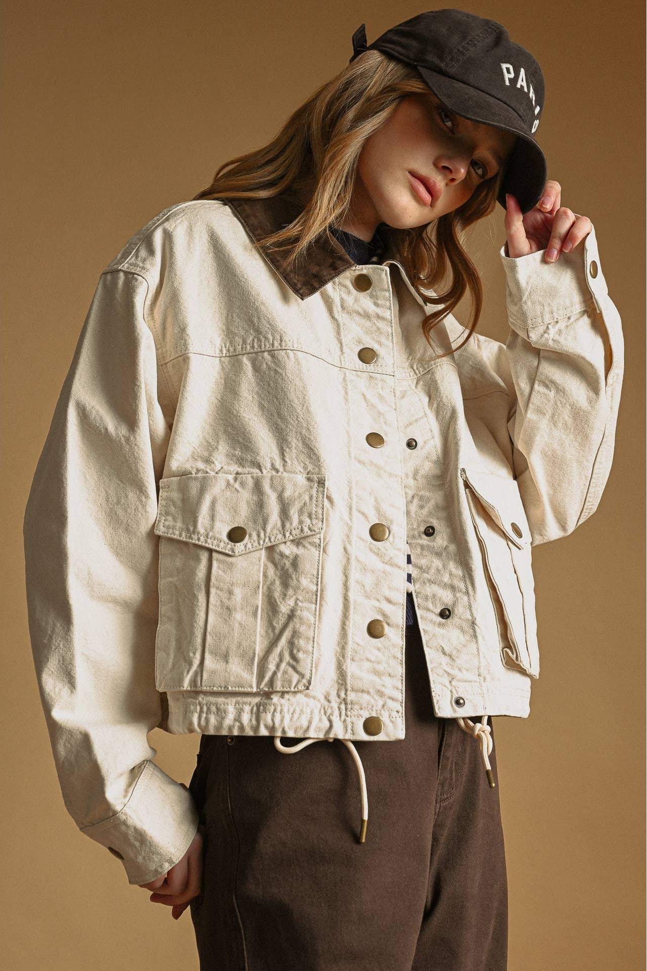 A woman wearing an ecru-colored cropped utility jacket with a dark brown collar, paired with dark brown pants and a black baseball cap, posed against a solid warm brown background.
