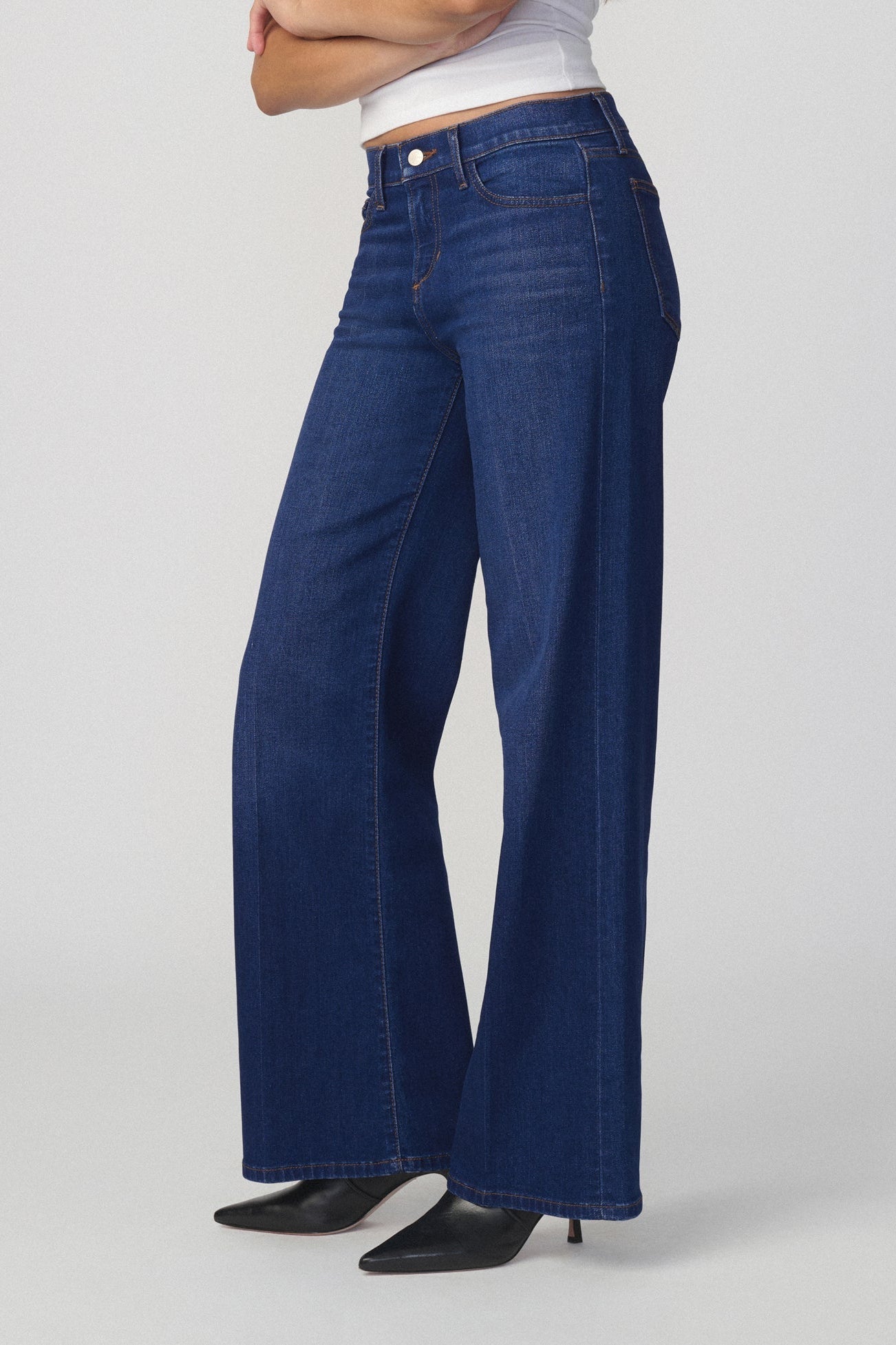 Side view of a person wearing dark blue wide-leg flare lou lou jeans in splendor color with a white cropped top and black pointed-toe heels, shown from the waist down against a light gray studio background.