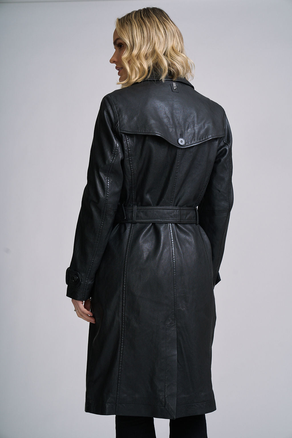 A woman shown from the back wearing a black leather trench coat with a matching belt, photographed against a plain light gray studio background.