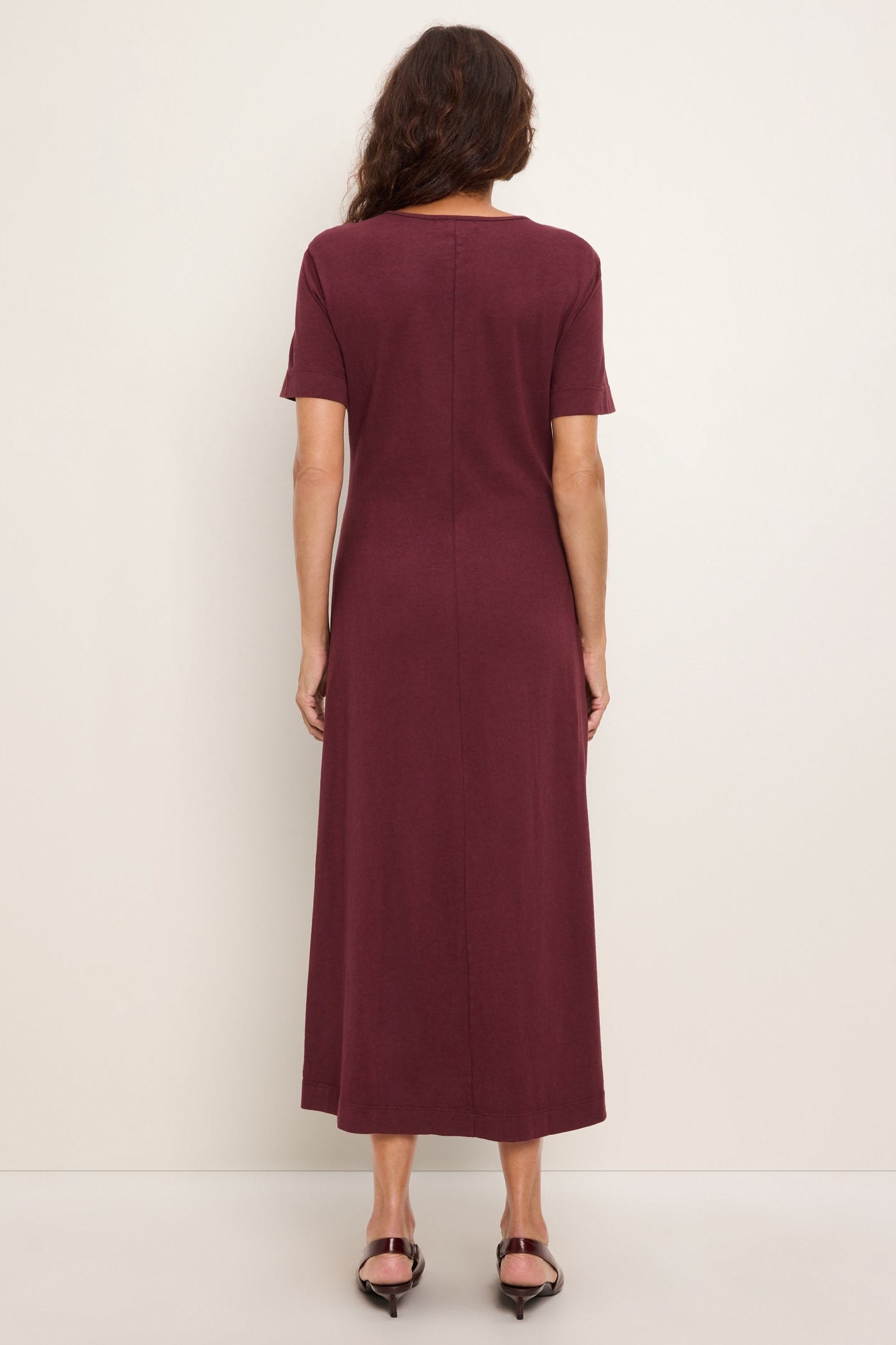 A woman viewed from the back wearing a short-sleeve, knee-length port royale colored column dress and black pointed-toe flats against a plain off-white studio background.
