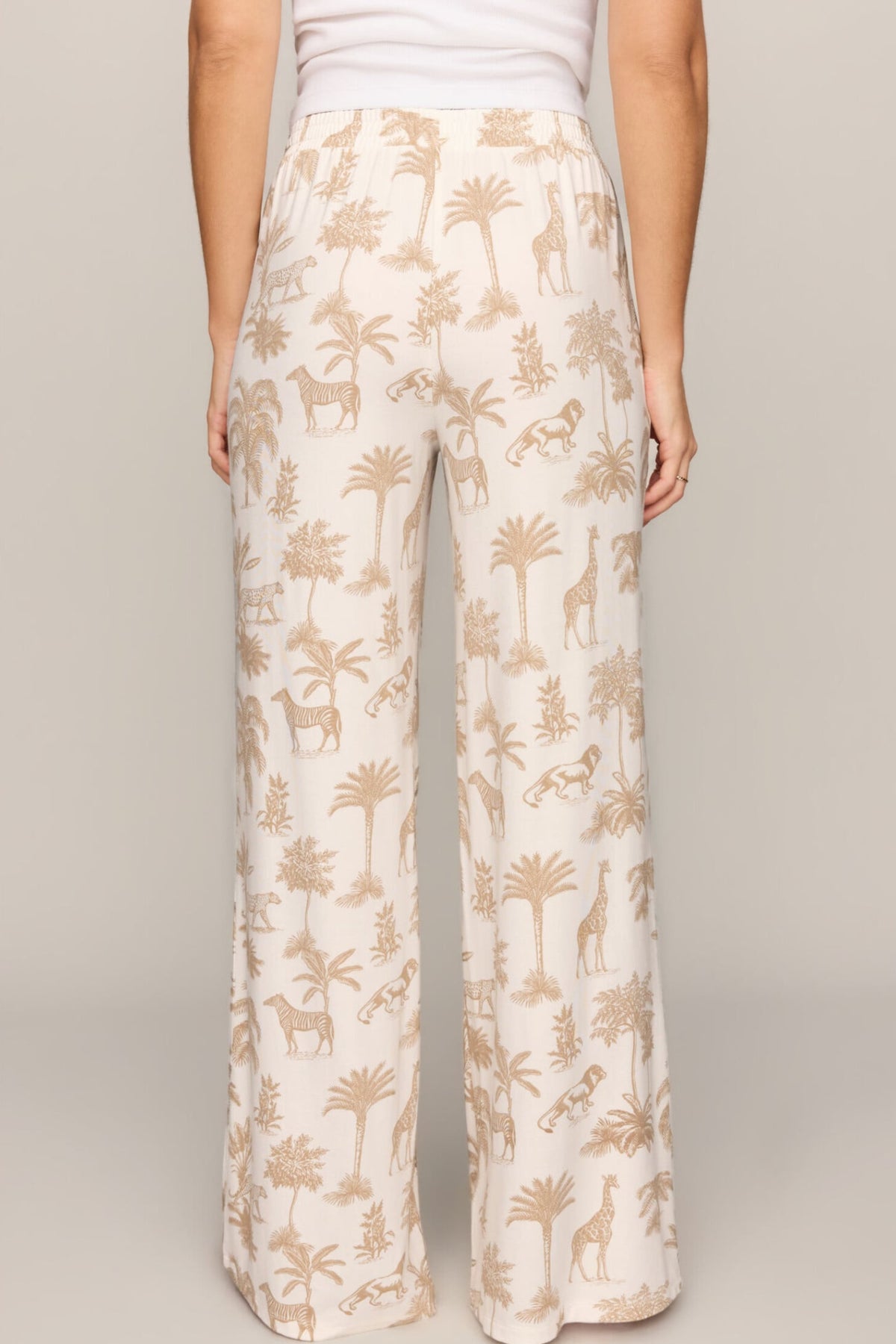 A person shown from the back wearing wide-leg pants with an allover safari-themed print of palm trees, giraffes, and zebras on a light cream background, paired with a white top against a neutral studio backdrop.