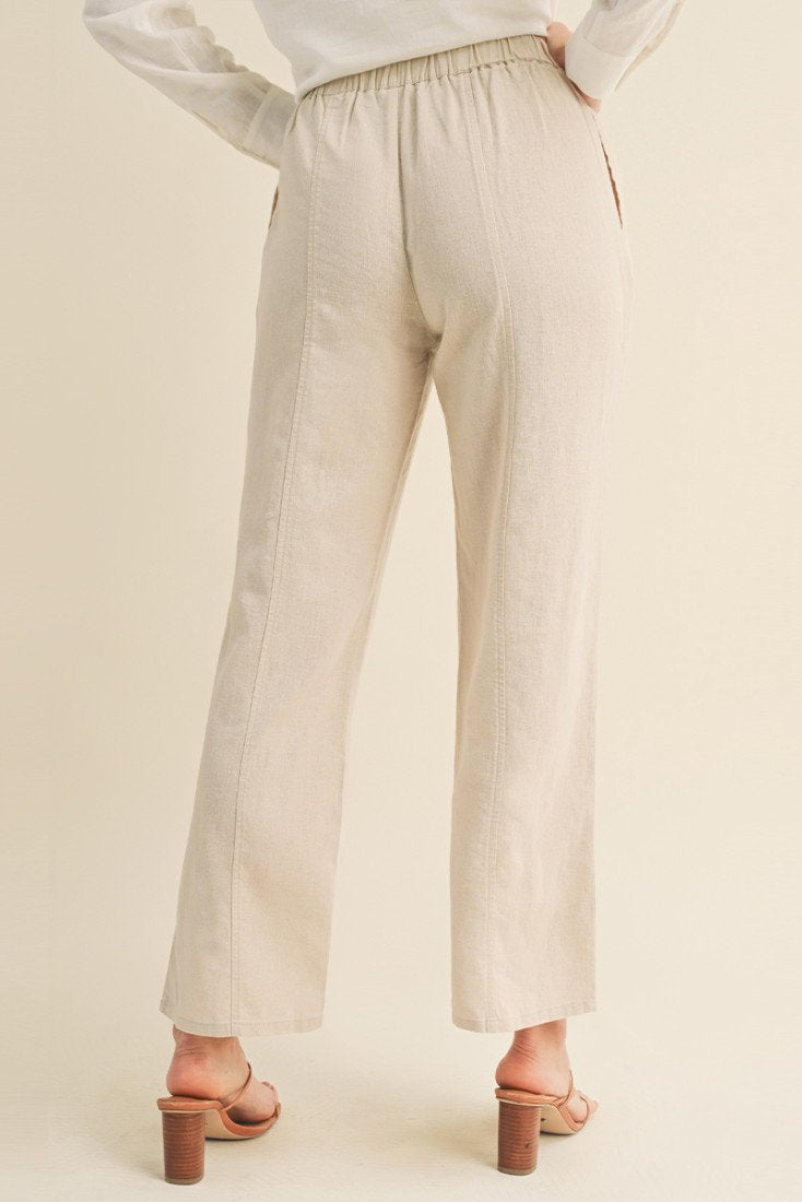 A person shown from the back wearing light beige khaki wide-leg cropped pants with an elastic waistband, paired with a white top and tan heeled slide sandals, against a solid warm beige studio background.
