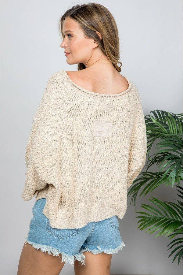 A woman wearing a chunky knit off-the-shoulder cropped patch sweater in oatmeal and light blue denim shorts, standing next to a green plant against a light gray wall.