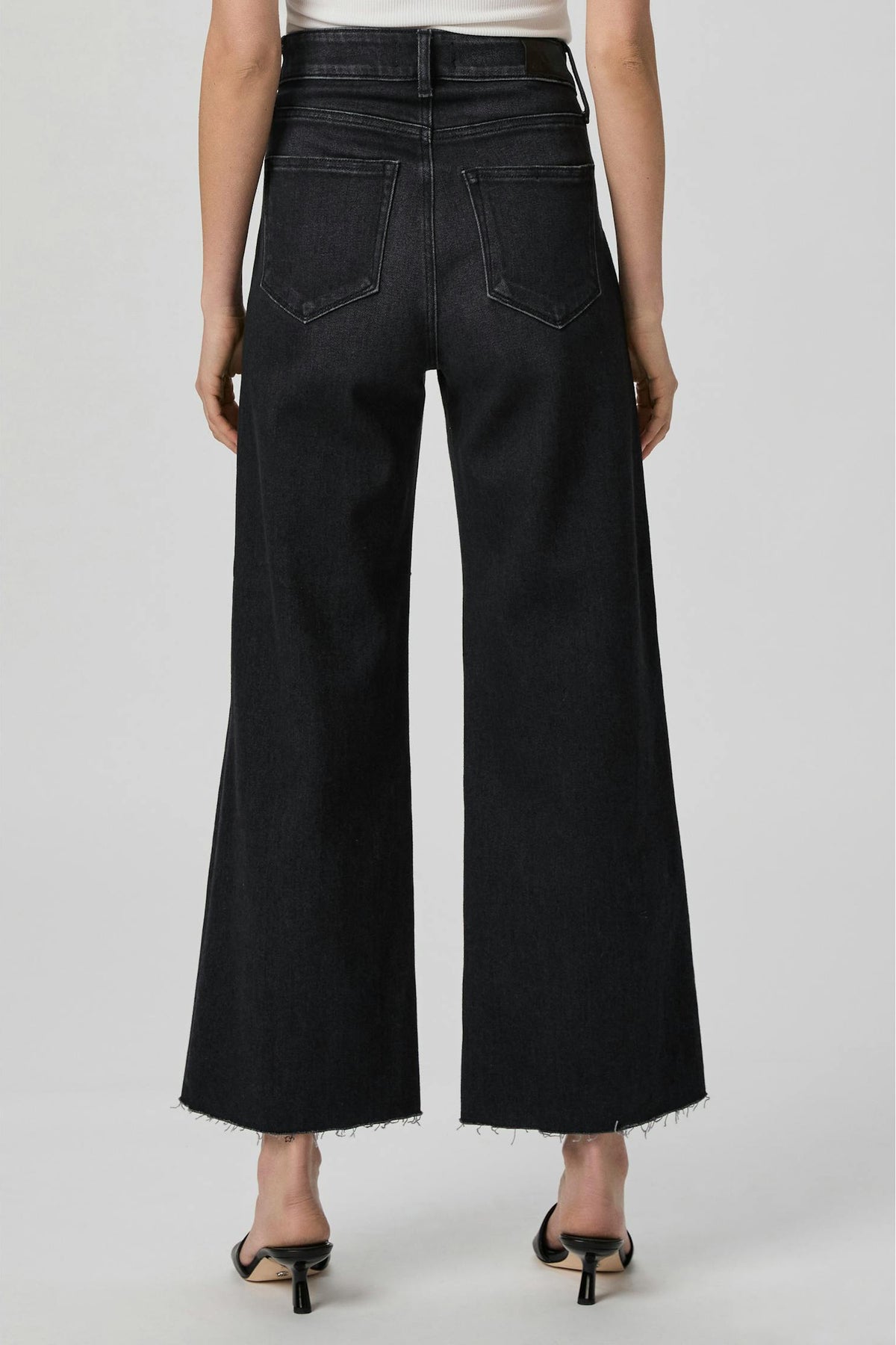 Rear view of a model wearing Anessa ink black wide-leg cropped jeans with a frayed hem and back pocket details, paired with black heeled sandals against a light gray studio background.