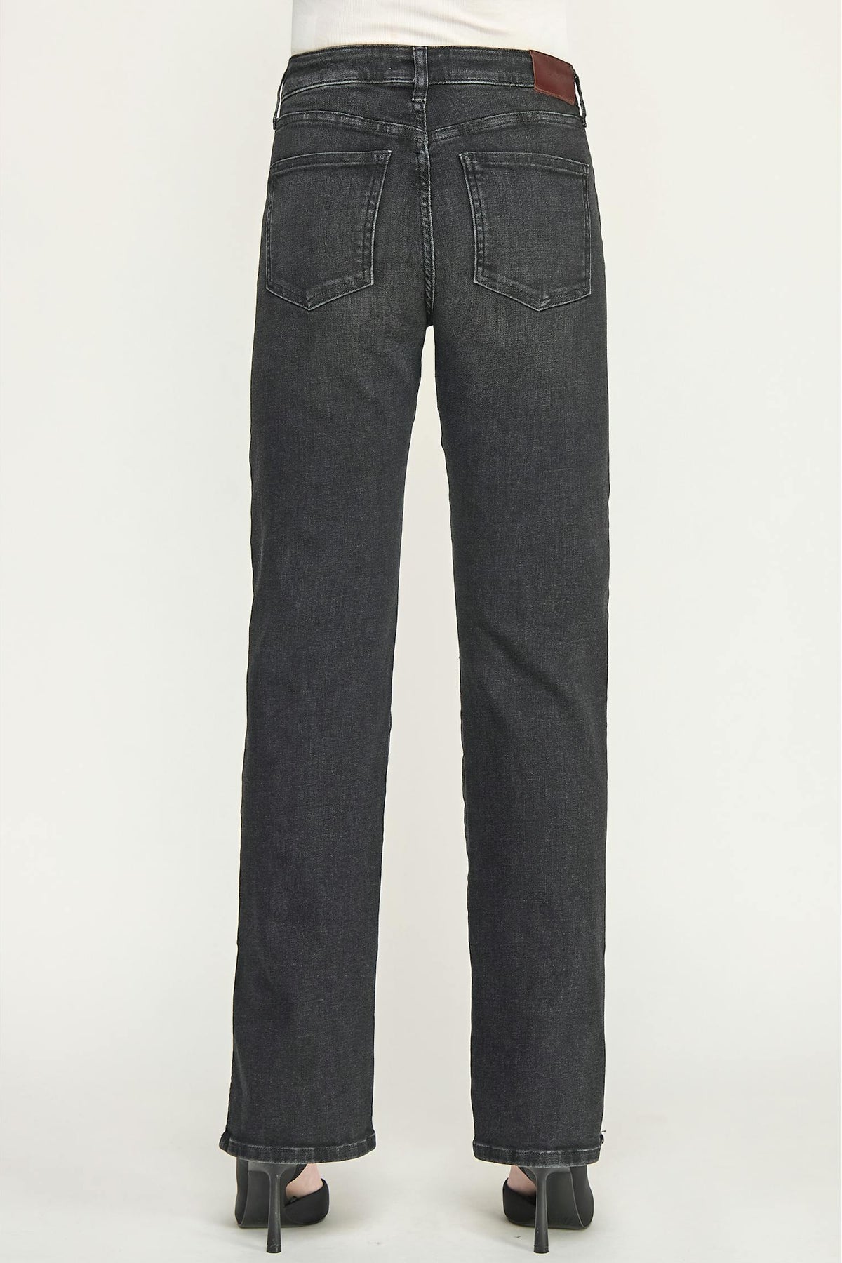 Rear view of a person wearing dark ash straight-leg cigarette jeans with a white top and black high heels, shown against a plain light gray studio background.