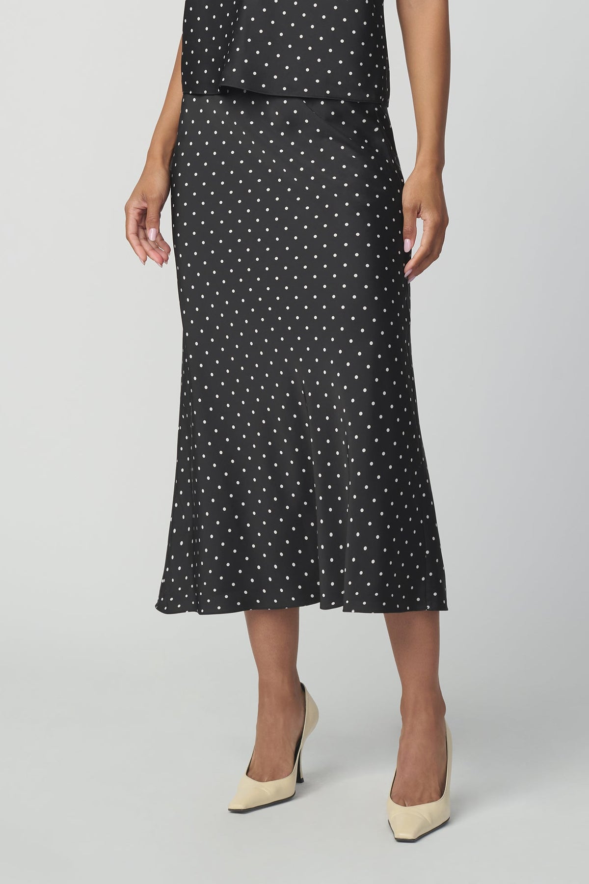 A model wearing a black midi skirt with a white polka-dot pattern, paired with beige pointed-toe heels, shown from the waist down against a light gray studio background.