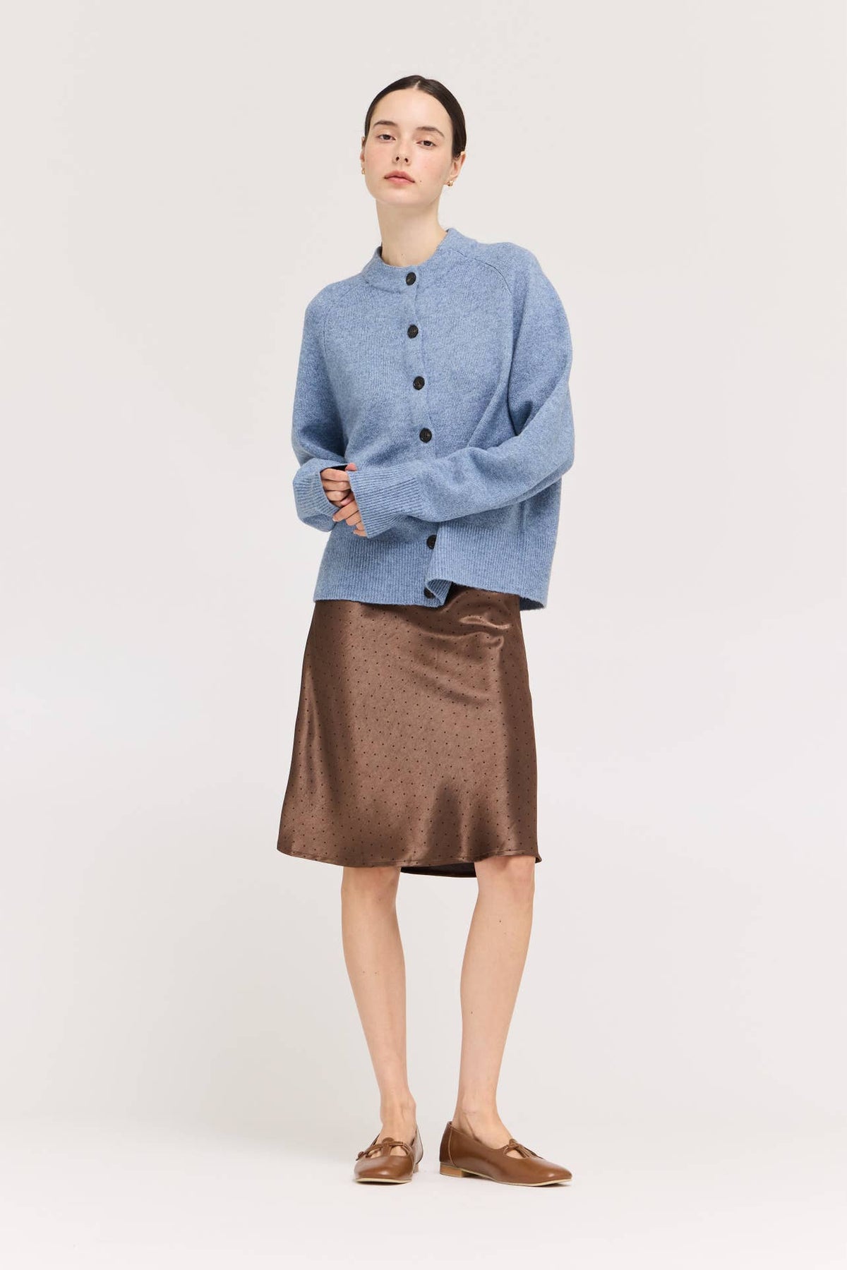 A model stands against a light gray studio background wearing a light blue cropped button-front cardigan, a chocolate brown satin A-line skirt with black polka dots, and brown loafers.