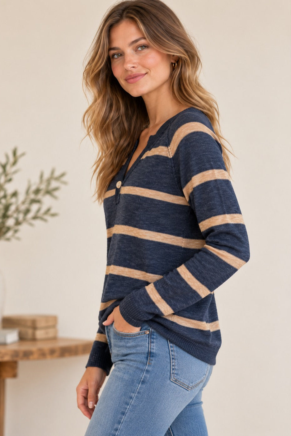 A woman wearing a navy midnight blue and beige striped long-sleeve henley sweater with light blue jeans, standing in a brightly lit indoor setting.