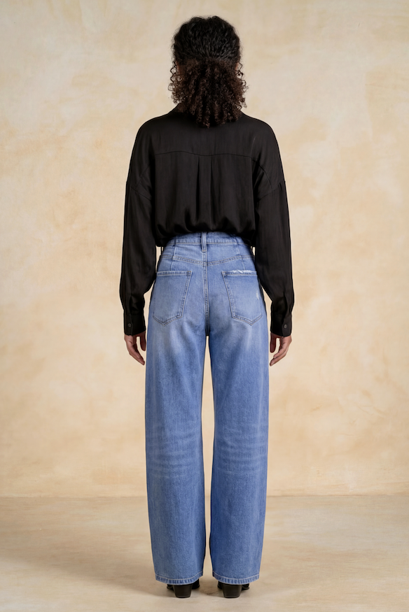A model viewed from the back wearing a black long-sleeve blouse and high rise barrel denim jeans, standing against a textured beige studio backdrop.
