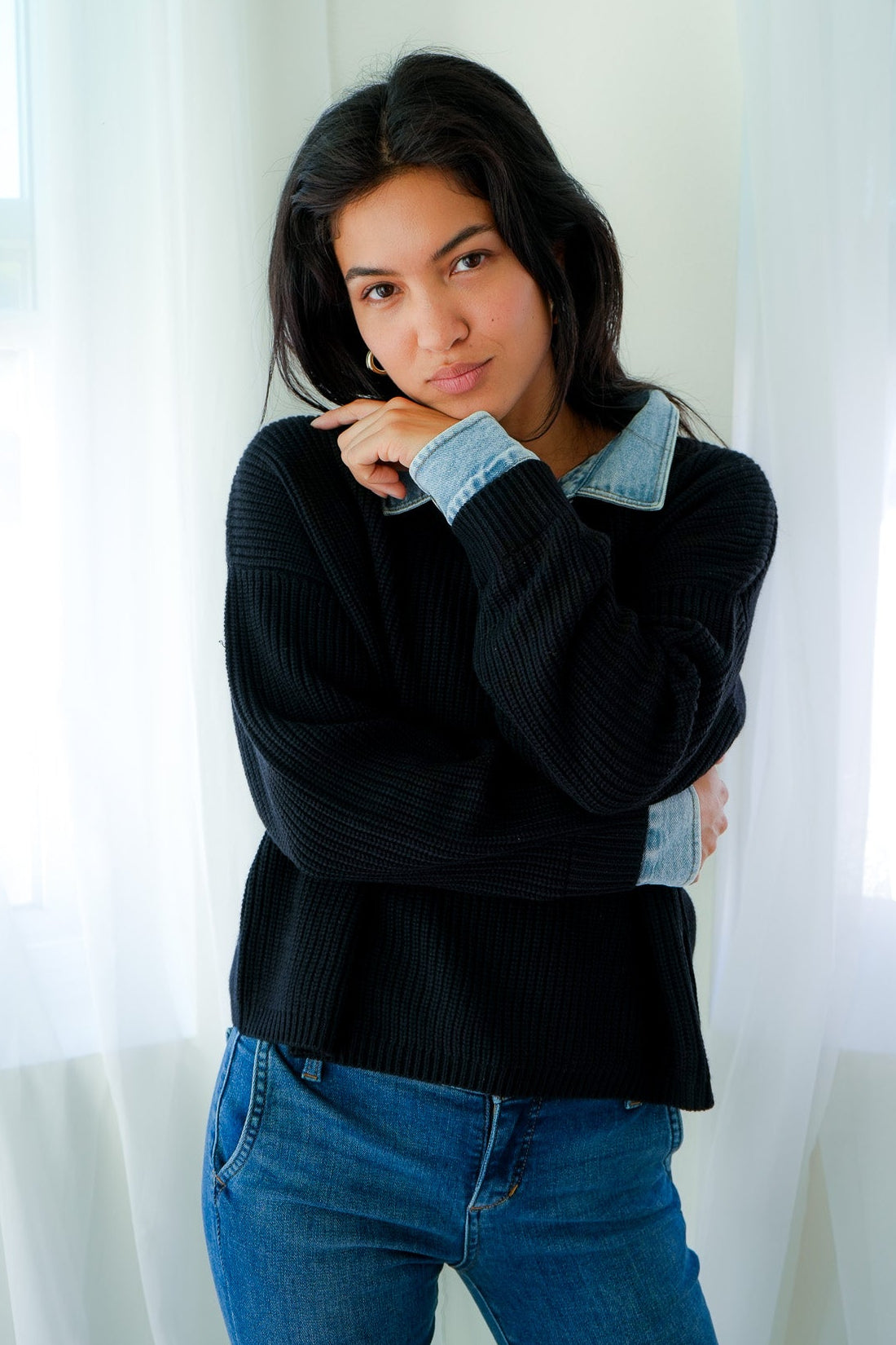 Woman wearing a black sweater and blue jeans standing in front of white curtains.
