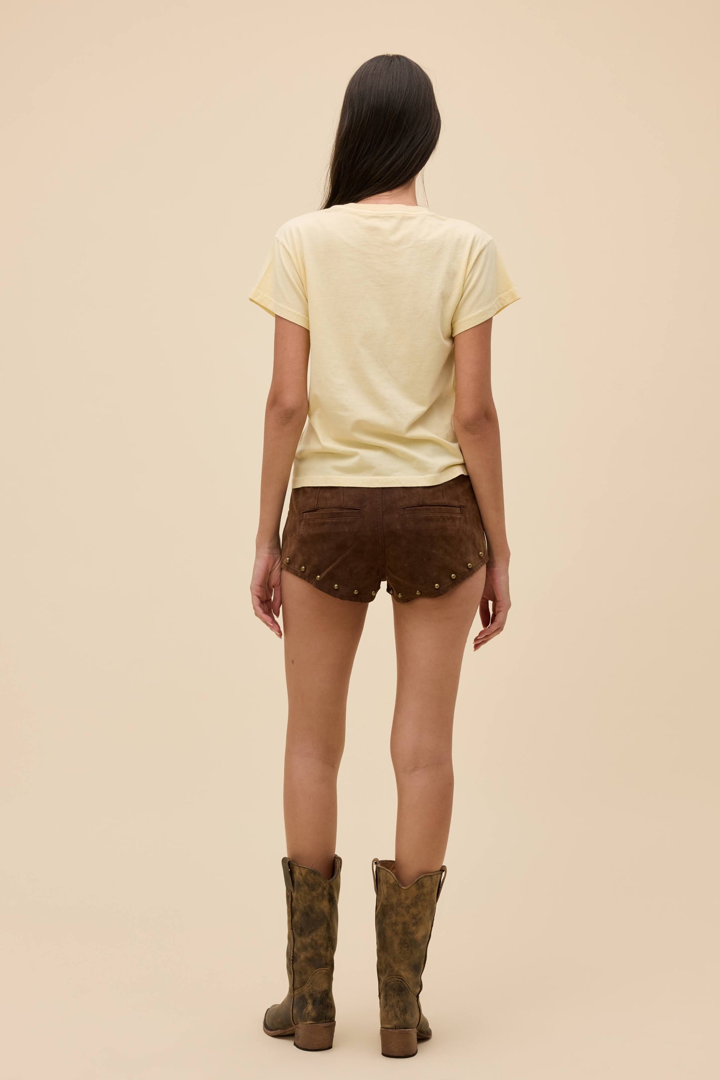 Back view of person wearing a sheer yellow top and brown shorts with studded details on a beige background