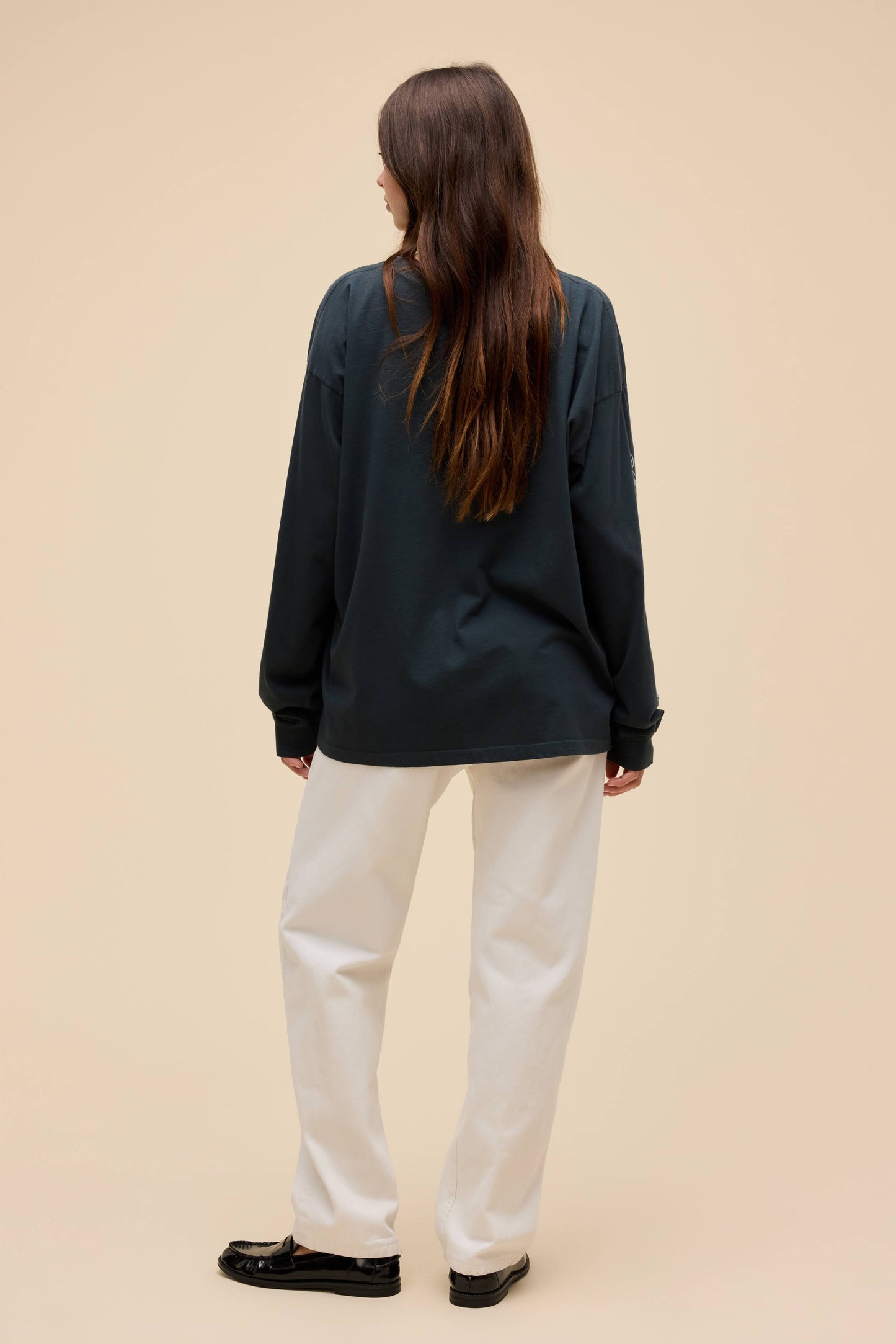 A woman shown from the back wearing an oversized vintage black long-sleeve top, off-white wide-leg trousers, and black loafers against a solid beige studio background.