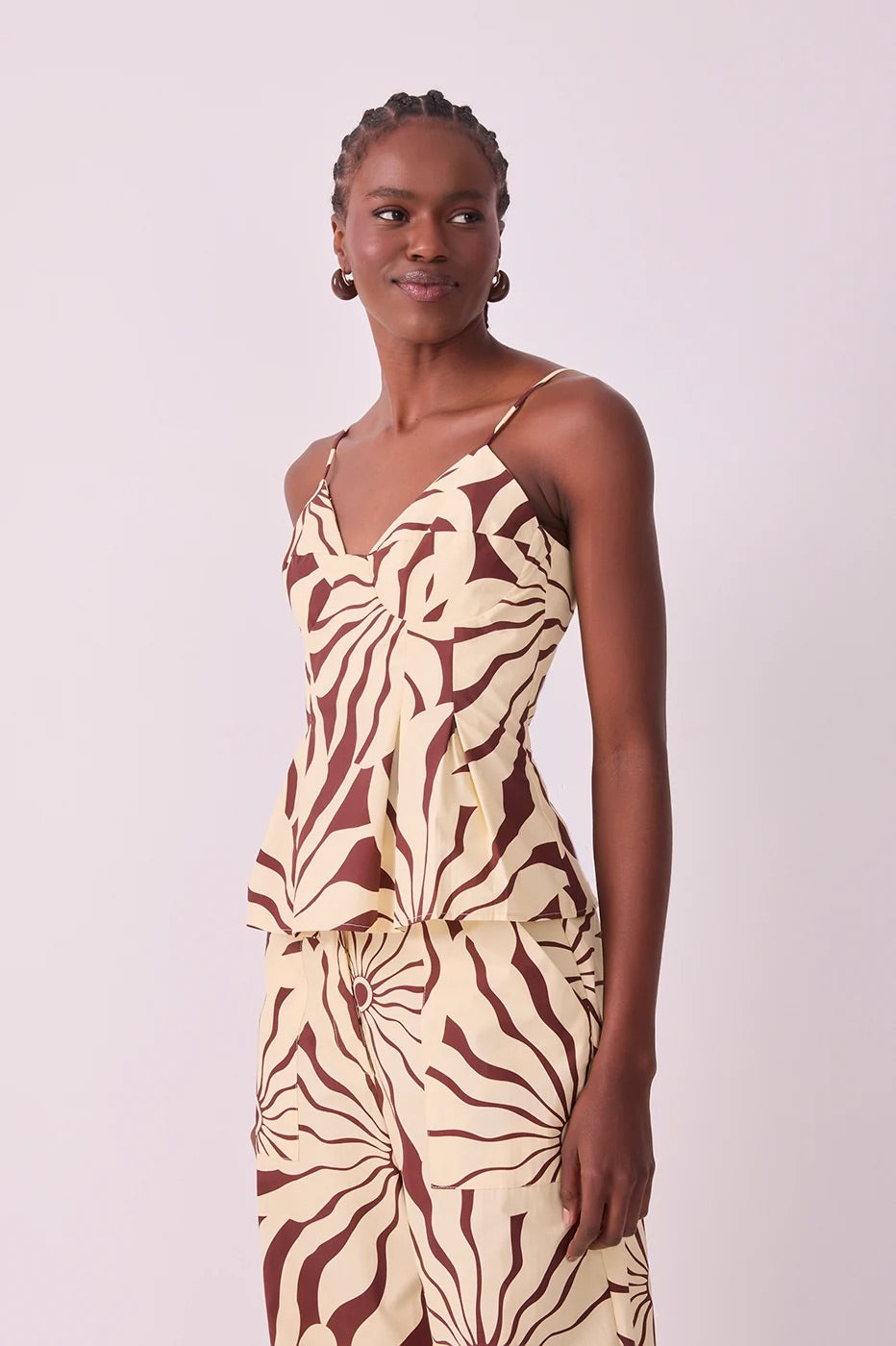 A woman modeling a sleeveless peplum-style top and matching pant set with a bold abstract alvorecer print in dark brown and cream, set against a plain light background.
