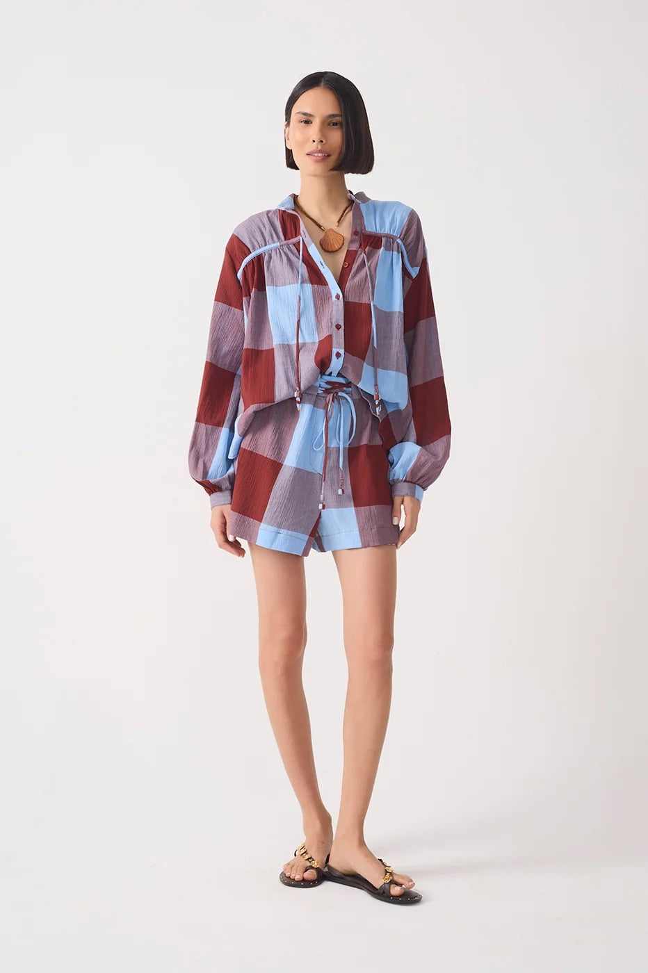 A model wearing a long-sleeve plaid shirt and matching shorts set in shades of red, blue, and white, styled with black sandals against a plain light gray studio background.