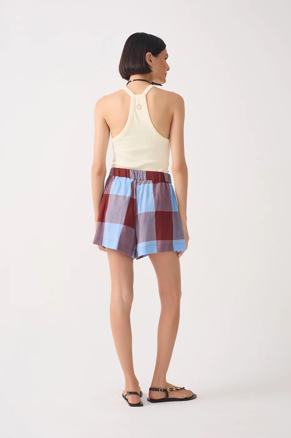 A model shown from the back wearing a cream halter top and a blue, burgundy, and gray plaid mini skirt, paired with brown sandals, against a plain white studio background.