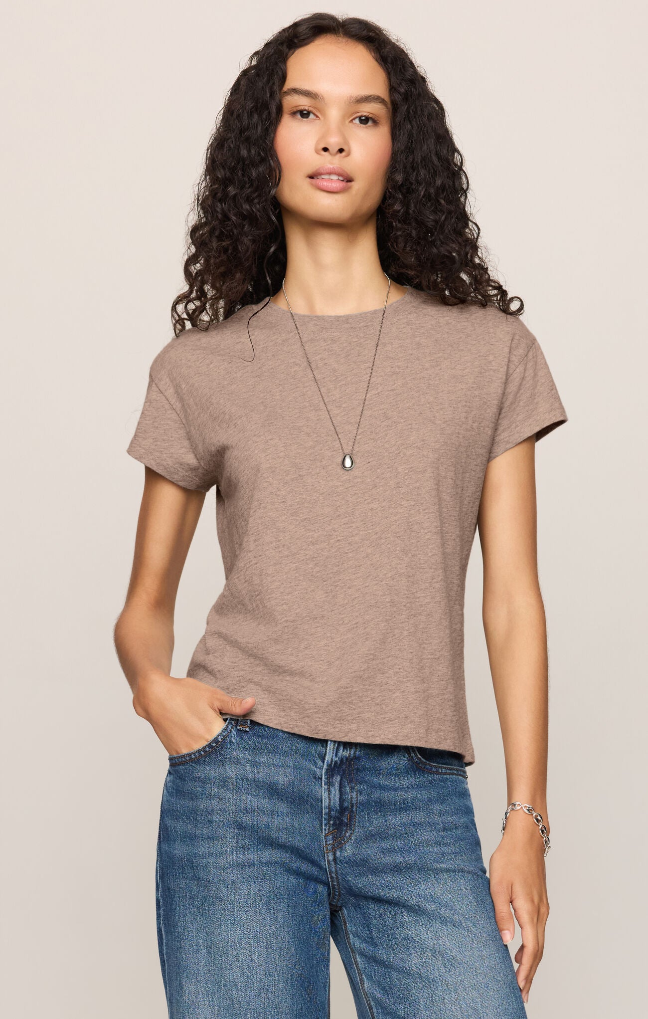 A woman with curly dark hair wearing a short-sleeve heather ash crewneck t-shirt paired with blue jeans, against a solid light beige background.