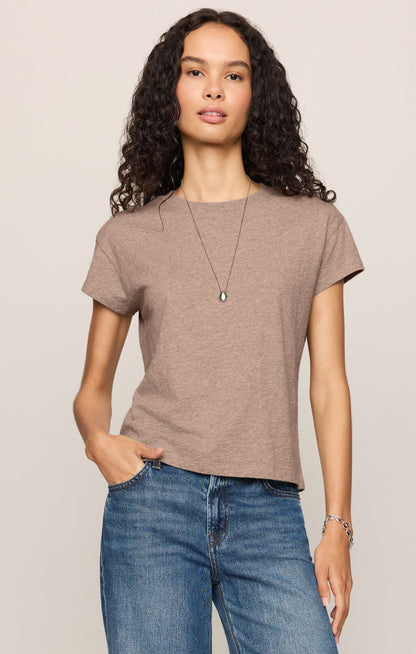 A woman with curly dark hair wearing a short-sleeve heather ash crewneck t-shirt paired with blue jeans, against a solid light beige background.