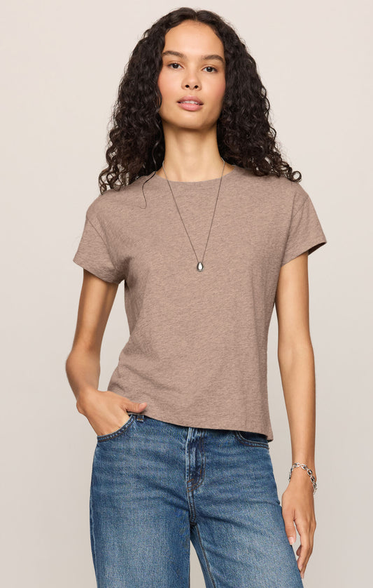 A woman with curly dark hair wearing a short-sleeve heather ash crewneck t-shirt paired with blue jeans, against a solid light beige background.