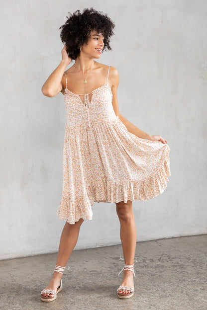 A woman modeling a natural & peach floral print spaghetti-strap dress with a tiered skirt, standing against a textured gray concrete wall.