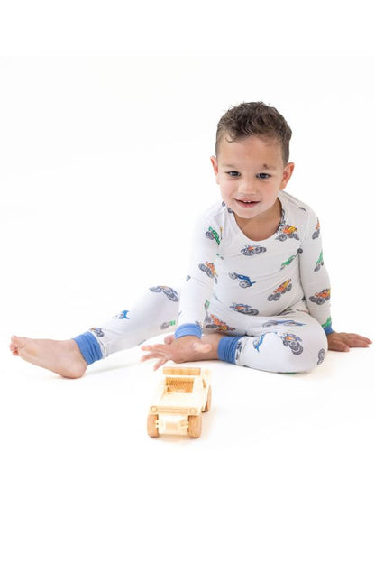 A young child sitting on a white studio floor wearing white long-sleeve pajama set with a colorful monster truck print and blue cuffs, interacting with a small wooden toy truck.