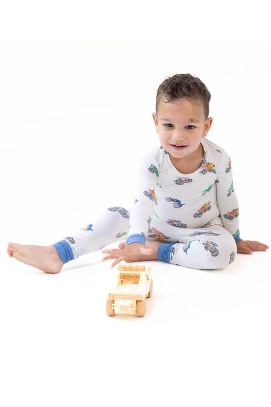 A young child sitting on a white studio floor wearing white long-sleeve pajama set with a colorful monster truck print and blue cuffs, interacting with a small wooden toy truck.