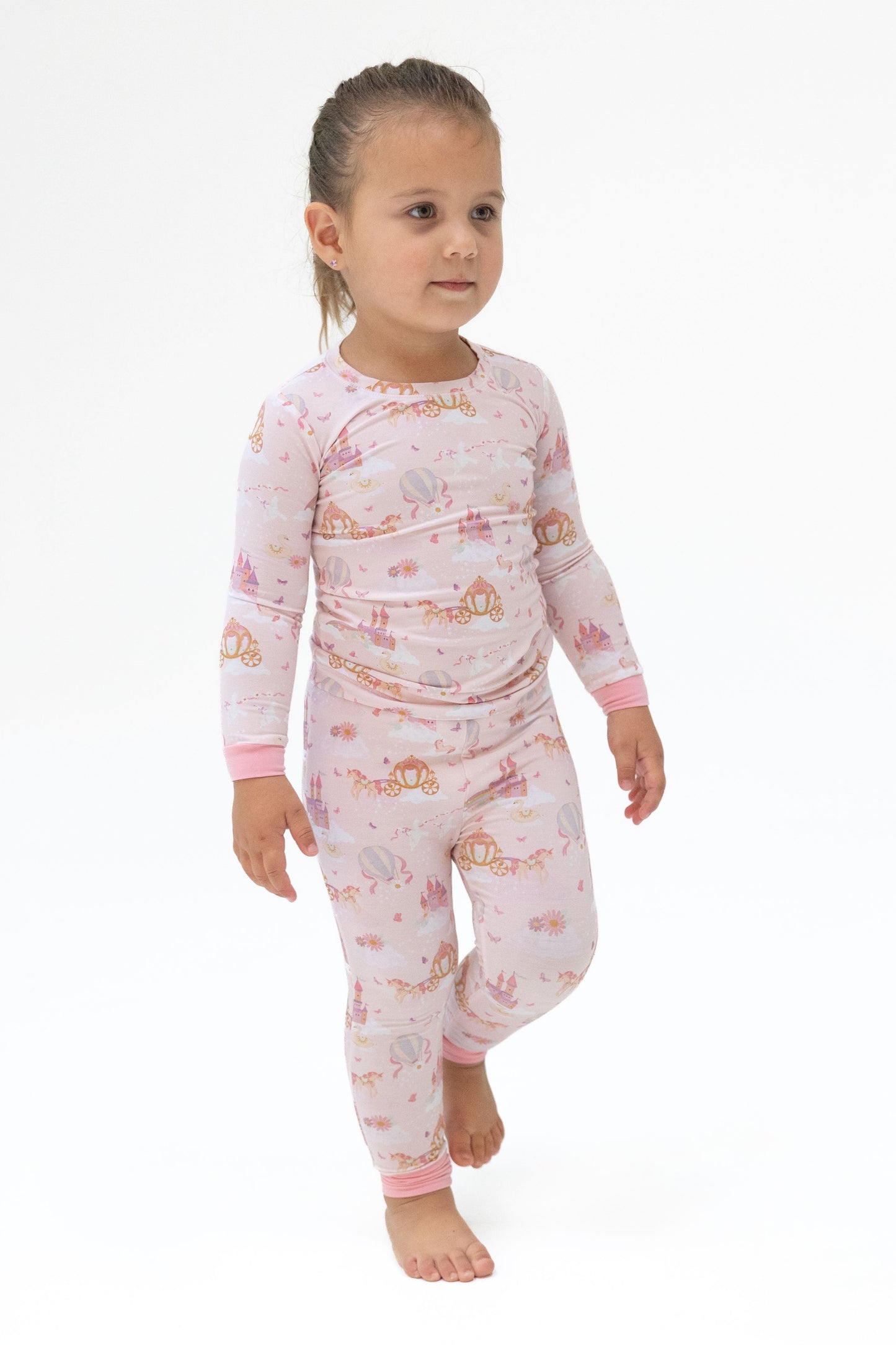 A young child wearing a light pink long-sleeve pajama set with a whimsical castle 'princess unicorns' print walking barefoot against a plain white background.