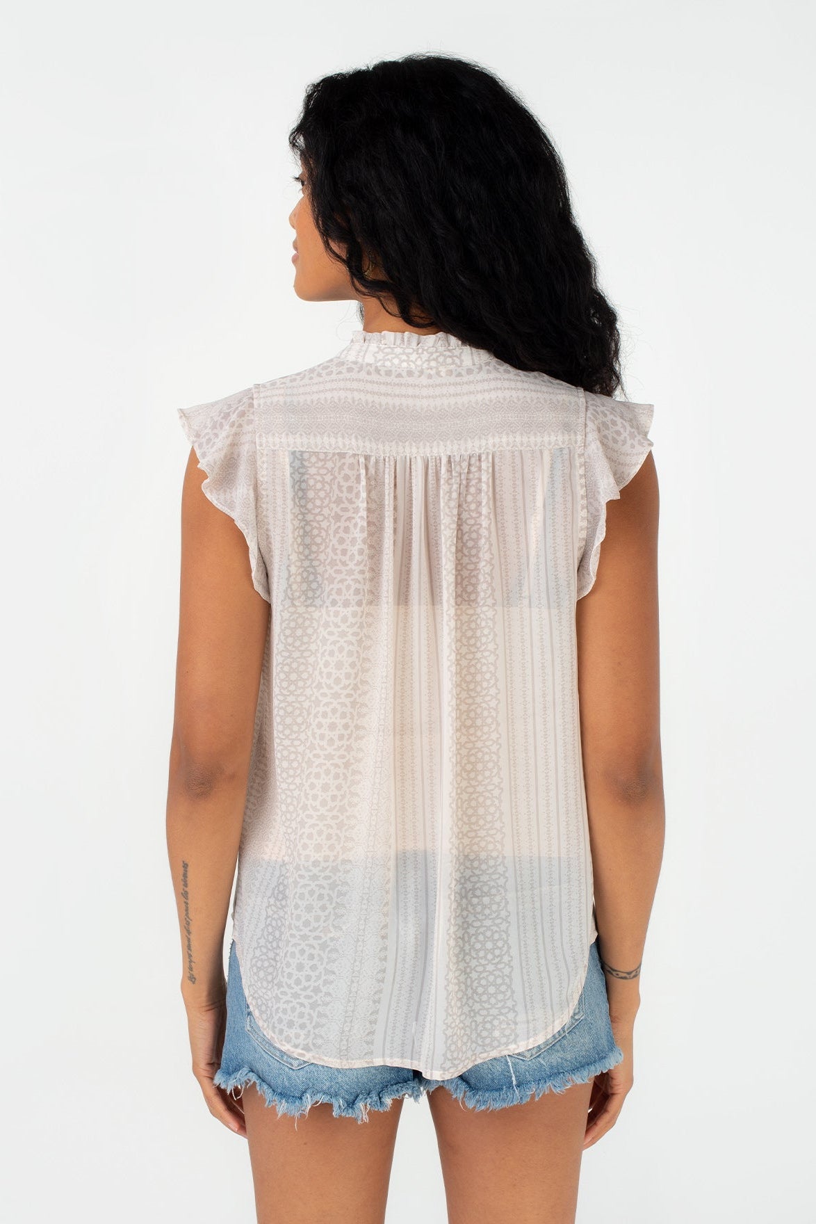 A woman shown from the back wearing a natural taupe sleeveless eyelet blouse with ruffled shoulders and denim shorts against a plain white background.