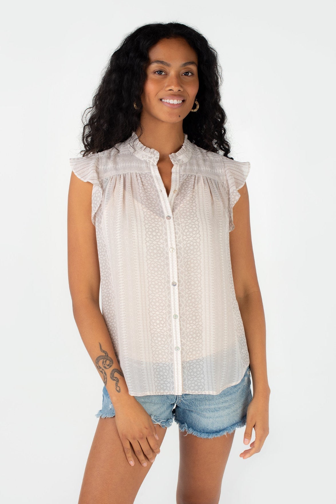 A woman modeling a sleeveless natural taupe button-up blouse with ruffled shoulders and a subtle dotted pattern, paired with denim shorts, against a plain white background.