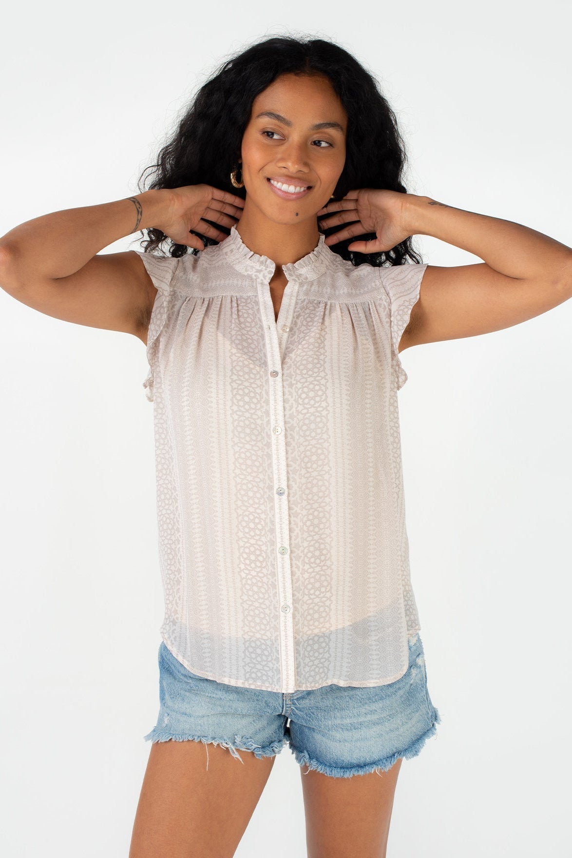 A woman modeling a ruffle sleeve natural taupe button-up blouse with a ruffled collar, paired with denim shorts, against a plain white background. 