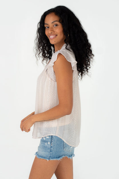 A woman with long, curly black hair is shown from a side-back angle wearing a natural taupe ruffle sleeve blouse with ruffle details and light blue denim shorts against a plain white background.