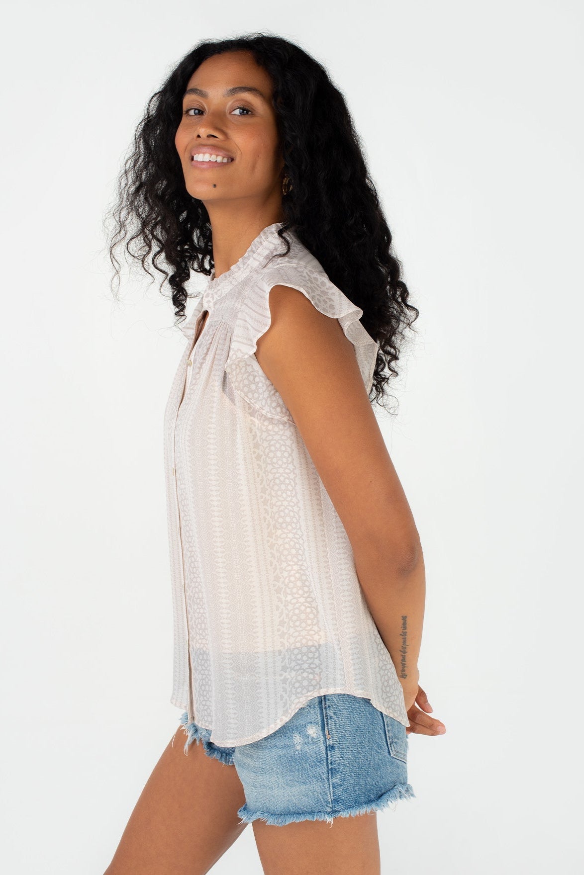 A woman wearing a natural taupe blouse with ruffled shoulders and a textured pattern, paired with denim shorts, photographed against a plain white background.