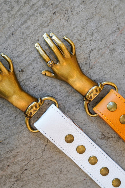 Gold hand-shaped straps with a white strap featuring gold studs on a textured surface