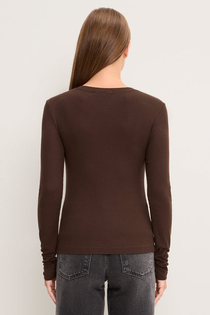 A woman viewed from the back wearing a fitted long-sleeve crew-neck top in molasses, paired with dark gray jeans, against a plain light gray studio background.