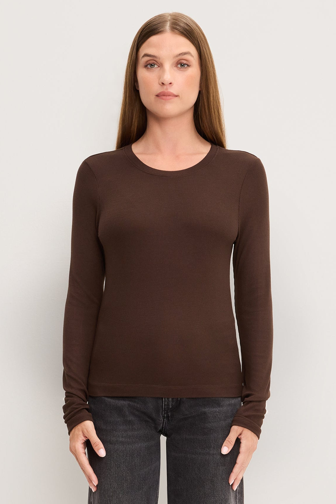 A woman modeling a molasses colored long-sleeve crewneck top paired with dark gray jeans, standing against a light gray studio background.