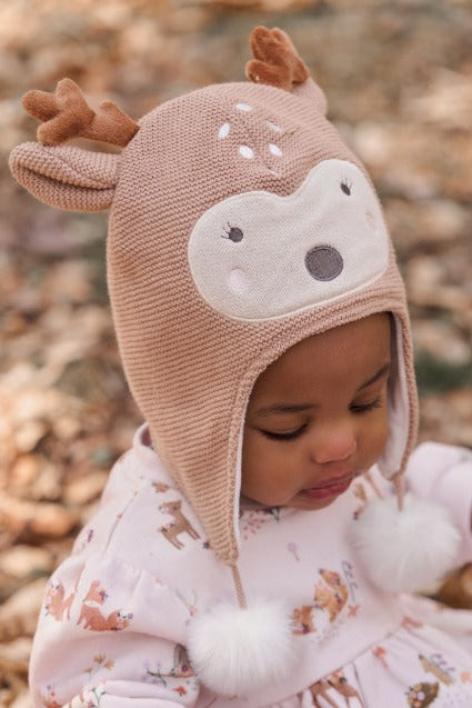 A baby wearing a knitted fawn deer-themed winter hat with ear flaps, faux fur pom-poms, and embroidered antlers, photographed outdoors on a background of fallen leaves.
