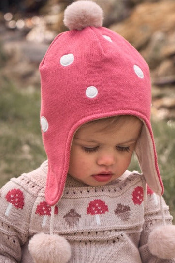 Child wearing a red mushroom knit hat with white polka dots and pom-poms, standing outdoors.