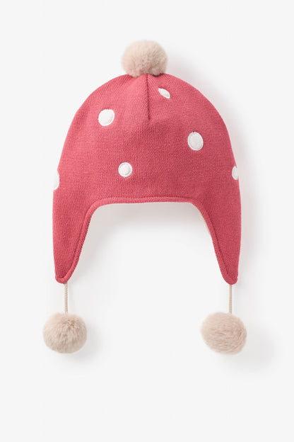 Red mushroom knit baby hat with white polka dots and pom-poms on a light beige background