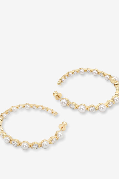 A pair of gold hoop earrings embellished with white pearls and small clear rhinestones, displayed on a light gray background.