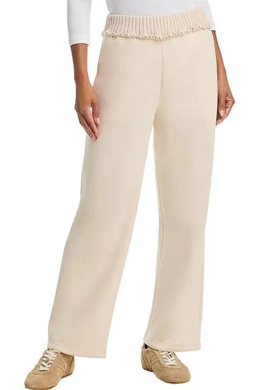 A person wearing  bone-colored wide-leg pants with a textured, ruffled  rib waistband, styled with a white long-sleeve top and beige sneakers against a plain white background.