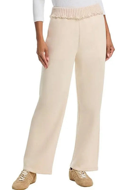 A person wearing  bone-colored wide-leg pants with a textured, ruffled  rib waistband, styled with a white long-sleeve top and beige sneakers against a plain white background.