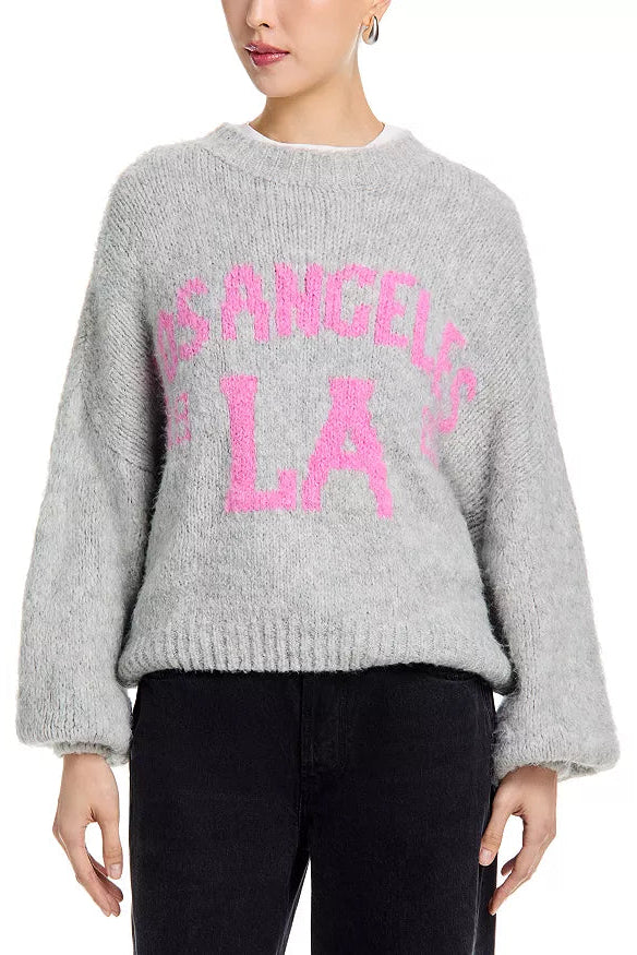 Woman wearing a light grey sweater with pink letters that say "Los Angeles LA" and black pants 