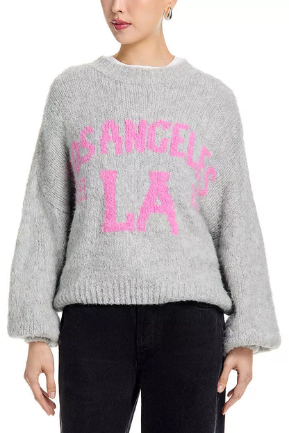 Woman wearing a light grey sweater with pink letters that say "Los Angeles LA" and black pants 