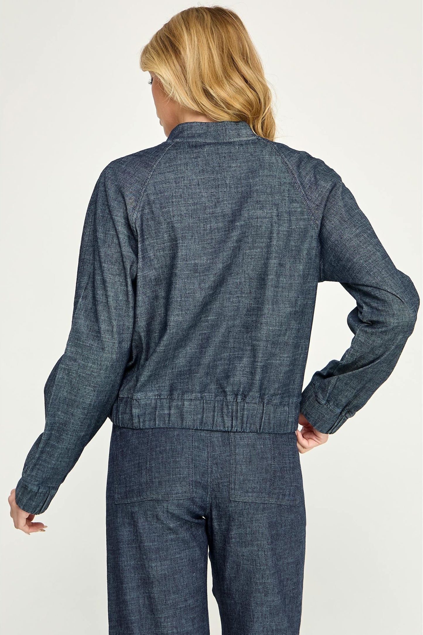 A woman shown from the back modeling a cropped, dark blue stargazer alex bomber denim jacket paired with matching denim pants against a light gray studio background.