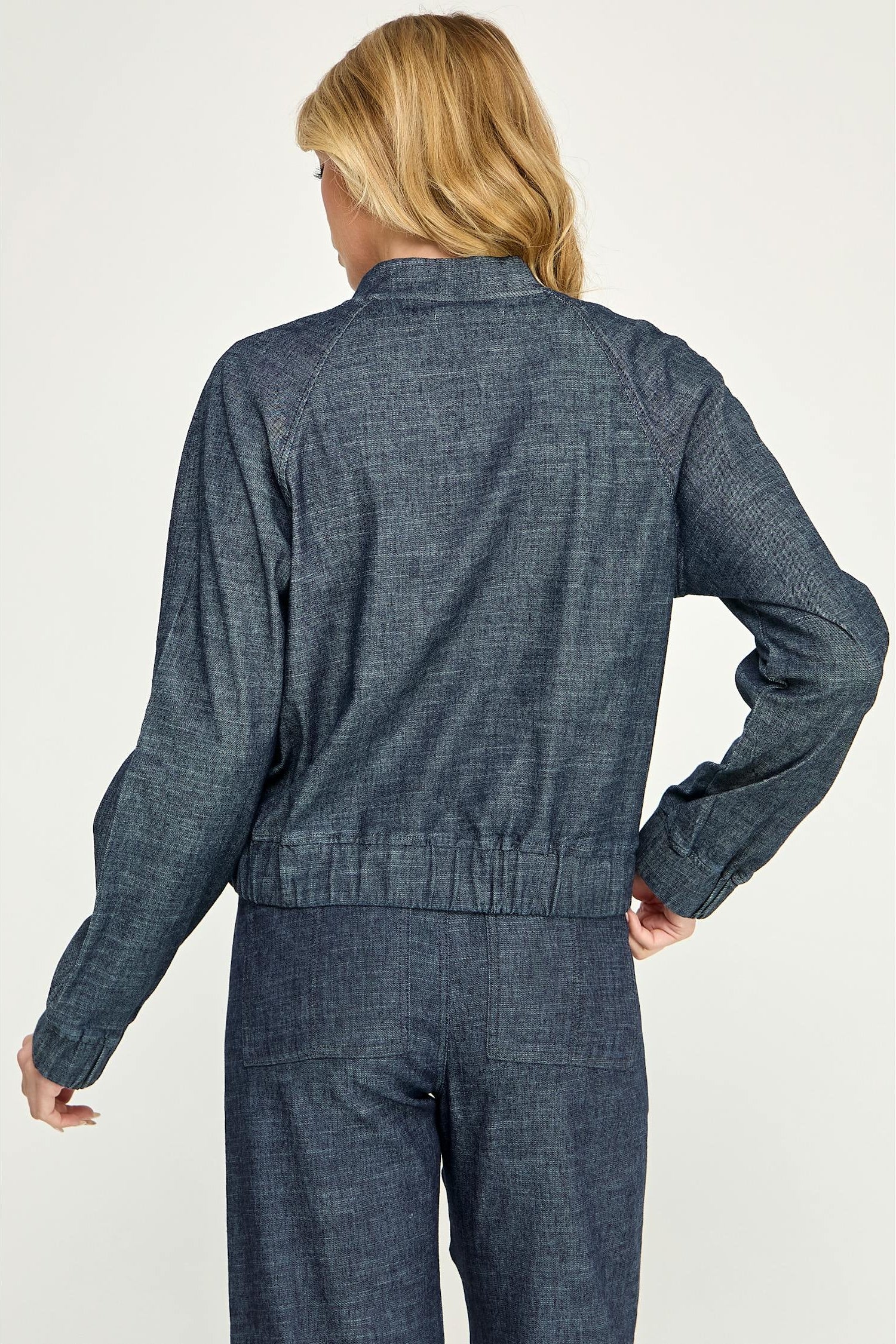 A woman shown from the back modeling a cropped, dark blue stargazer alex bomber denim jacket paired with matching denim pants against a light gray studio background.